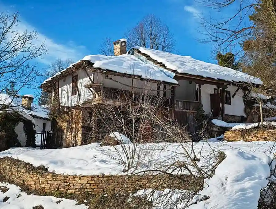 Weathered rustic mountain cabin covered in snow, surrounded by leafless trees and a bright blue sky.