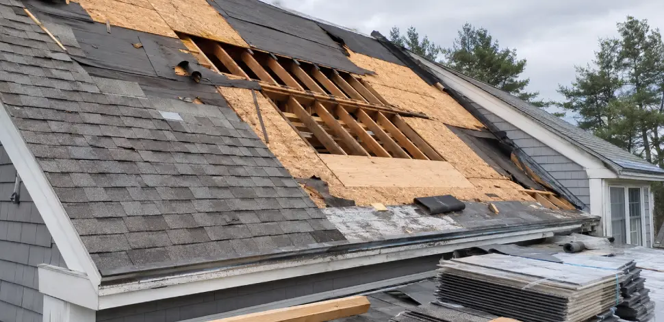 Storm-damaged roof undergoing reconstruction with exposed sheathing, damaged underlayment, and staged roofing materials.
